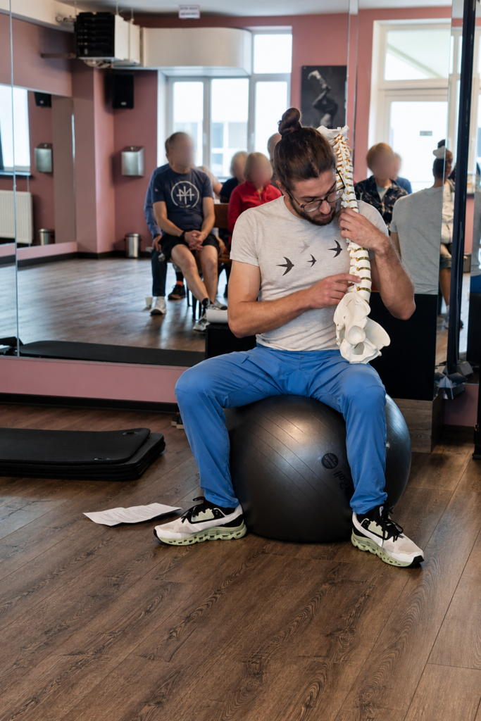 A fitness instructor holds a spine model while explaining anatomy to participants in a mirrored exercise studio.